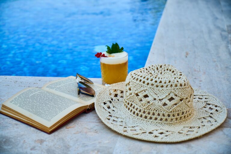 beige straw hat book sunglasses and drink beside pool. Best books in June 2023: recommendations