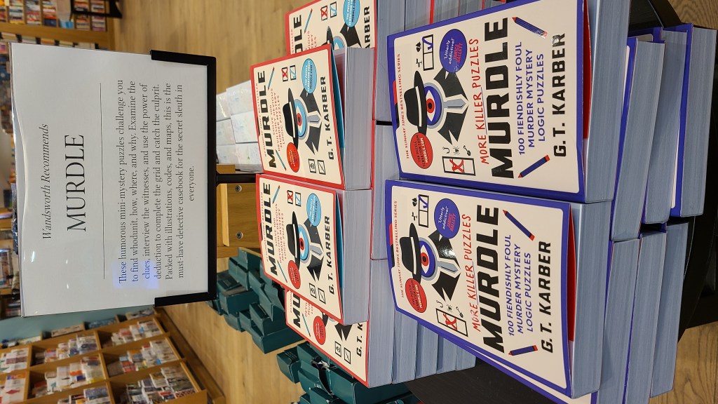 Murdle at Waterstones. Sign on book stand says: "These humorous mini-mystery puzzles challenge you to find whodunit, how, where, and why. Examine the clues, interview the witnesses, and use the power of deduction to complete the grid and catch the culprit. Packed with illustrations, codes, and maps, this is the must-have detective casebook for the secret sleuth in everyone.
