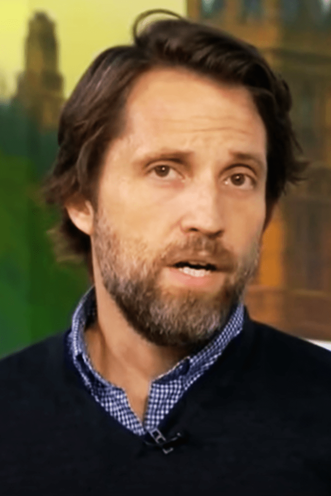 A Day in the Life of Abed Salama author Nathan Thrall with brown hair and beard wears a blue chequered shirt and navy jumper. Parliament is in the background.