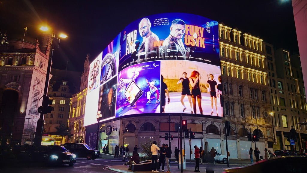 Piccadilly Circus is featured in Harry Potter. A bustling city scene at night showing a vibrant display of digital billboards and advertisements, including a boxing match promotion. The urban environment is lively with pedestrians and traffic under the illuminated signs.