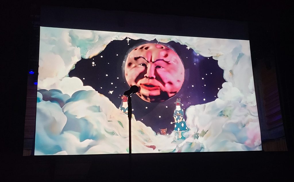 The image shows a stage with a microphone stand in front of a large screen displaying an animated graphic. The graphic features a stylized image of the moon with a human face, surrounded by clouds and stars. The moon's expression is calm, with eyes closed, set against a dark night sky. Below the moon, small figures appear on clouds, including one holding a lantern, enhancing a dreamlike, whimsical atmosphere for Rough Trade Books event