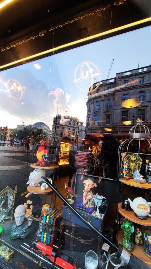 Harry Potter souvenir shop near Charing Cross Road. A themed store interior seen through a window, filled with assorted memorabilia and fantastical elements reflecting a popular magical universe. The reflection of a bustling city street outside contrasts with the enchanting items inside.