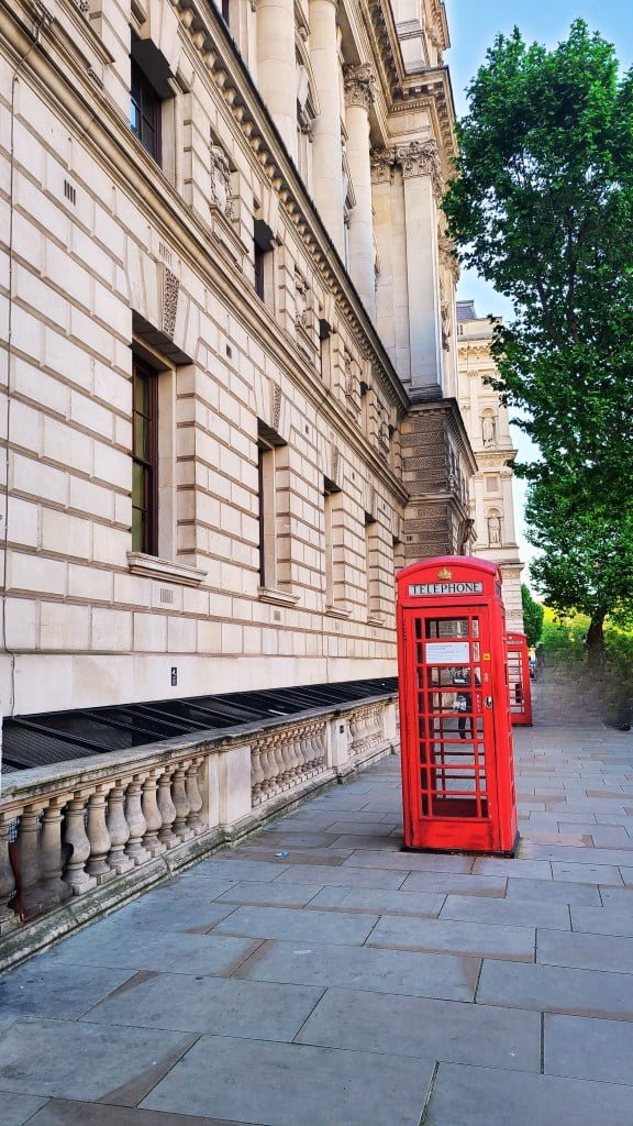 Red post box. A classic London street view featuring an iconic red telephone booth set against the backdrop of a grand white stone building with ornate architectural details and lush greenery, evoking a sense of traditional British urban scenery.