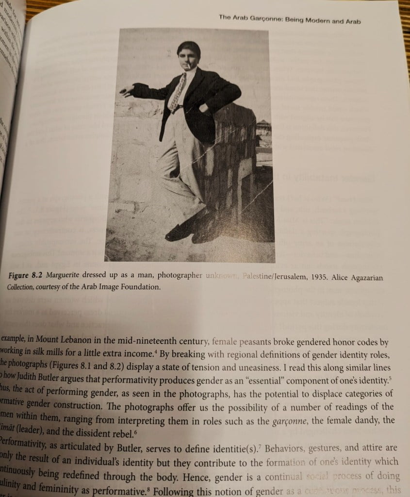 Books by Palestinian women authors. A vintage black and white photo depicting a person posing with one hand on their hip and the other extended outward. They are dressed in early 20th-century Western masculine attire, with a blazer, tie, and trousers, standing confidently on a stone block outdoors. The backdrop is nondescript, with a light sky and hints of a rural landscape. This image is labeled "Figure 8.2 Marguerite dressed up as a man, photographer unknown, Palestine/Jerusalem, 1935. Alice Agazarian Collection, courtesy of the Arab Image Foundation." The page is from a book discussing the concept of gender identity and performativity in the context of Middle Eastern history, highlighting the fluidity and construction of gender roles.
