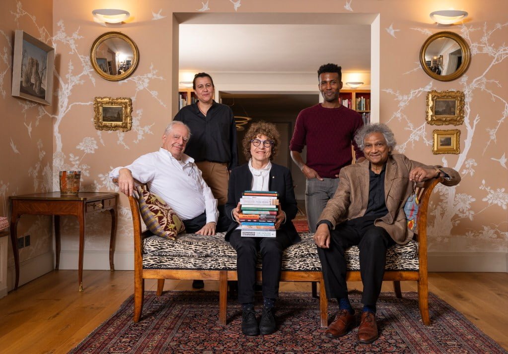 International Booker Prize 2024 judges holding books featured in the longlist. They sit on top of a leopard print wooden seat in a orange and white flowery room the mirrors and paintings on the wall.