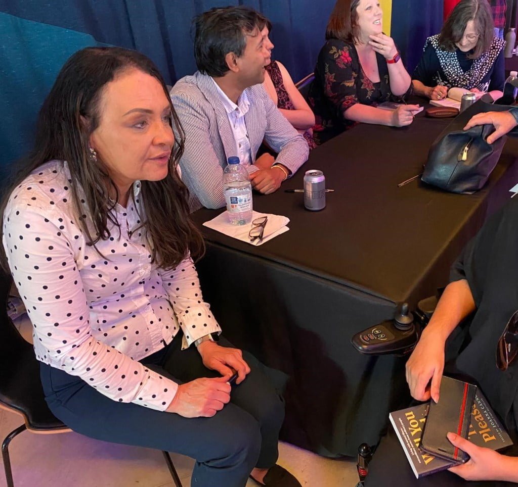 'Will You Read This, Please?' editor Joanna Cannon speaks to Suswati Basu. Cannon is wearing a black and white polka dot shirt whilst speaking to a woman on a wheelchair holding the book. The panellist appears behind her engaged in conversation.