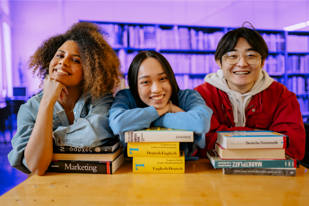 Three joyful students from diverse backgrounds lean on a stack of books in a library, smiling warmly at the camera. Libraries Change Lives Week on integral role in UK