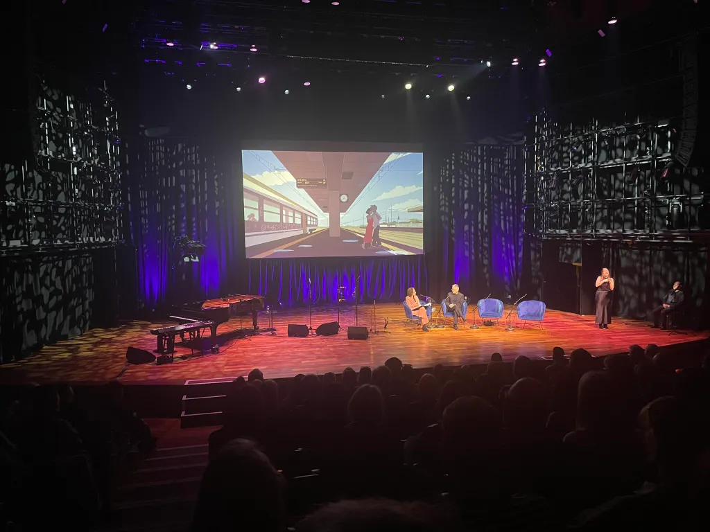 An event stage with an animated backdrop of a person standing by a train, Samira Ahmed and Kazuo Ishiguro sitting in blue chairs in the foreground, and a sign language expert addressing the audience on the right. The scene is set within a dark auditorium, illuminated by stage lights, with a captivated audience in attendance.